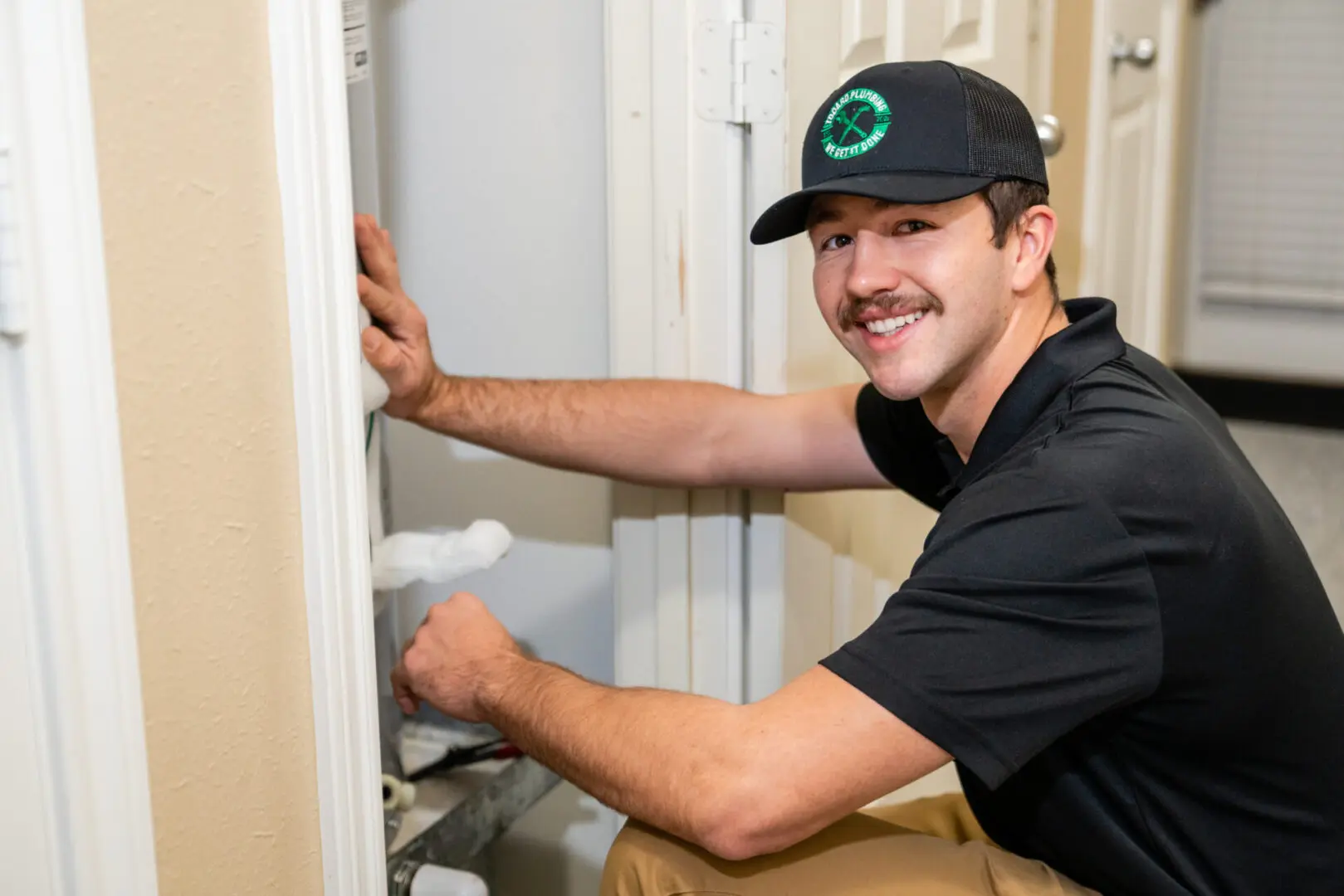 Todaro Plumber in a black polo shirt and black hat with a green logo, smiling while finishing up a tankless water heater installation in a closet inside a home in Bryan College Station Tx - Todaro Plumbing